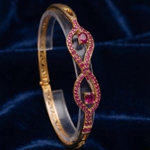 May include: A gold hinged bangle bracelet featuring a central infinity-shaped design adorned with numerous small round pinkish-red stones and two larger oval-cut pinkish-red stones set within the center of each loop of the infinity pattern.