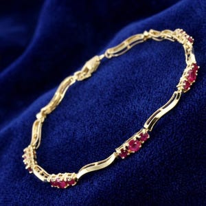 May include: A gold-toned bracelet with a series of oval-shaped, red gemstones. The bracelet has a delicate, wavy design. The gemstones are evenly spaced along the length of the bracelet. The background is a deep blue.