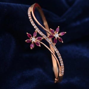 May include: A gold-toned hinged bangle bracelet featuring a curved, open-work design adorned with small, clear pavé-set stones. Two floral motifs are positioned along the band, each crafted with six marquise-cut deep red stones and a central clear stone.