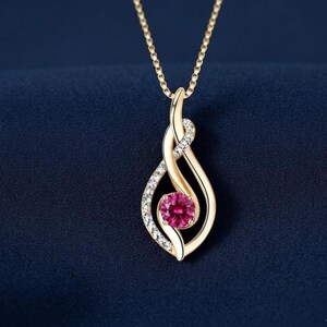 May include: A gold pendant necklace featuring a central round-cut pink gemstone set within a swirling, teardrop-shaped frame. One side of the polished gold frame is accented with a row of small, clear, sparkling stones on a delicate gold chain.