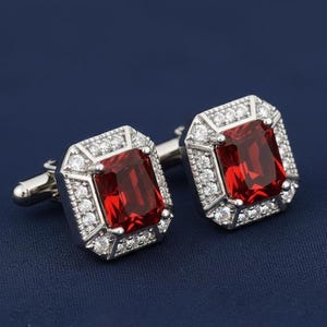 May include: A pair of silver cufflinks with a rectangular, deep red gemstone center. The gemstones are surrounded by a halo of smaller, clear stones. The cufflinks are set against a dark blue background.