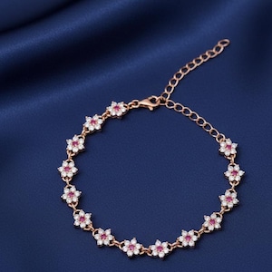 Op de afbeelding: Roségouden armband met een bloemmotief. Elke bloem heeft een rood hart en is omgeven door kleine, heldere stenen. De armband wordt gepresenteerd op een donkerblauwe achtergrond.