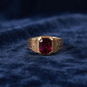 May include: A gold ring featuring a rectangular, deep red gemstone set in a prominent, structured metal band. The setting has a stepped, geometric design that frames the central stone, which displays a rich, saturated crimson hue and facets.