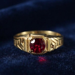Puede incluir: Un anillo de oro con una piedra preciosa redonda de color rojo intenso engastada en un bisel cuadrado. La banda presenta un patrón geométrico texturizado con líneas repetitivas en forma de galón a ambos lados del engaste central.