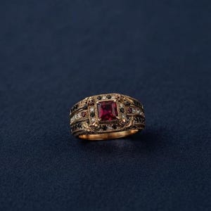 Könnte beinhalten: Ein goldener Ring mit einem zentralen quadratischen roten Edelstein, umgeben von einem Kranz aus kleinen weißen und schwarzen Steinen. Die Ringschiene ist mit detaillierten Blattgravuren sowie weiteren roten und schwarzen Steinen verziert.