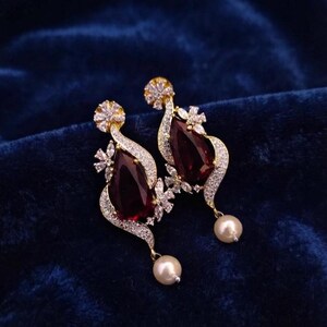 May include: A pair of gold-tone earrings featuring a large, teardrop-shaped dark red stone surrounded by small clear crystals. The design includes a curved, crystal-encrusted frame and a single white pearl dangling from the bottom of each earring.