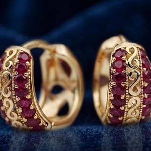 Peut inclure: Une paire de créoles dorées présentant une rangée centrale de pierres rondes rouge foncé, encadrée par deux bandes décoratives avec un motif en S et des détails perlés le long des bords de la monture métallique.