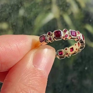 Könnte beinhalten: Ein goldfarbener Ring mit einer Reihe von quadratischen und runden roten Edelsteinen. Die Edelsteine sind in einem sich wiederholenden Muster um das Band angeordnet. Der Ring wird vor einem verschwommenen grünen Hintergrund gehalten.