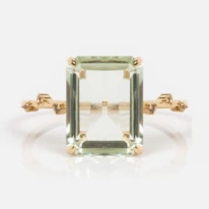 Könnte beinhalten: Ein goldener Ring mit einem großen, rechteckigen, hellgrünen Edelstein. Der Edelstein ist in einer goldenen Fassung gefasst. Das Band hat ein strukturiertes Design mit kleinen goldenen Akzenten auf jeder Seite des Edelsteins.