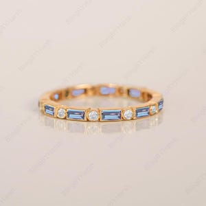 Könnte beinhalten: Ein goldfarbener Ring mit abwechselnd rechteckigen blauen und runden weißen Edelsteinen. Der Ring hat ein durchgehendes Banddesign mit mehreren gefassten Edelsteinen. Der Ring wird auf einer hellen Oberfläche gezeigt.