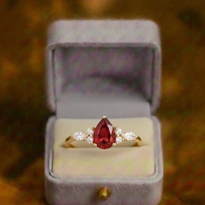 Könnte beinhalten: Ein goldener Ring mit einem tropfenförmigen, tiefroten Edelstein als Mittelpunkt, flankiert von kleineren, weißen Edelsteinen. Der Ring wird in einer grauen Samtringbox vor einem verschwommenen, goldbraunen Hintergrund präsentiert.