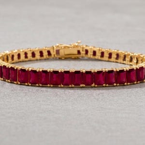 May include: Gold-toned bracelet featuring a row of rectangular, deep red gemstones. The bracelet has a secure clasp and is set on a neutral gray surface. A piece of jewelry suitable for special occasions.