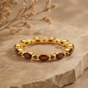 May include: A gold-colored ring with alternating oval and round red gemstones. The ring is set on a light brown, textured surface, with a sprig of dried flowers in the background. The ring is a piece of jewelry.