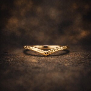 Könnte beinhalten: Ein V-förmiger Goldring mit einem eingravierten Blatt- und Rankenmuster entlang der Ringschiene. Das Metall hat eine polierte Oberfläche und das Design bildet eine ausgeprägte Chevron-Form, die zu einem Mittelpunkt zuläuft und botanische Details zeigt.