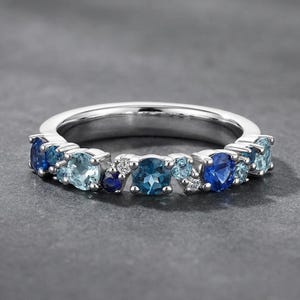 Könnte beinhalten: Ein silberner Ring mit einer Anordnung blauer Edelsteine. Der Ring zeigt eine Vielfalt an Blautönen, von hellem Aquamarin bis zu tiefem Saphir, durchsetzt mit kleinen, klaren Akzentsteinen. Die Edelsteine sind in einem polierten Silberband gefasst.