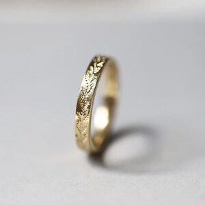 Könnte beinhalten: Ein goldener Ring mit einem filigranen floralen Muster, das um das Band geätzt ist. Der Ring besteht aus einem glänzenden, gelben Metall und ist vor einem weichen, neutralen Hintergrund platziert. Das Design des Rings zeigt kleine Blätter und Ranken.