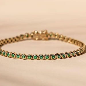 Könnte beinhalten: Ein goldfarbenes Tennisarmband mit einer Reihe von runden, leuchtend grünen Edelsteinen. Das Armband ist vor einem neutralen Hintergrund platziert, der das Funkeln der Edelsteine und den warmen Ton des Metalls hervorhebt.