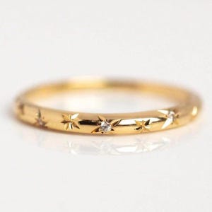 Könnte beinhalten: Ein goldener Ring mit einem Band, das mit sternförmigen Gravuren und kleinen, klaren Edelsteinen verziert ist. Der Ring hat eine polierte Oberfläche und ist vor einem weißen Hintergrund platziert. Dieses Schmuckstück ist für den täglichen Gebrauch geeignet.