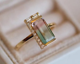 May include: A gold ring featuring a rectangular, multi-colored gemstone with pink, green, and clear sections. The ring has a delicate band and is accented with small, clear stones and rectangular accents.