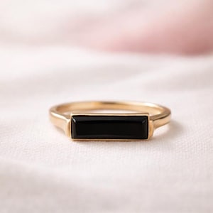 Könnte beinhalten: Goldfarbener Ring mit einem rechteckigen schwarzen Stein in der Mitte. Der Ring liegt auf einer weißen Oberfläche, mit einem zarten rosa Hintergrund. Schlichtes, elegantes Design.