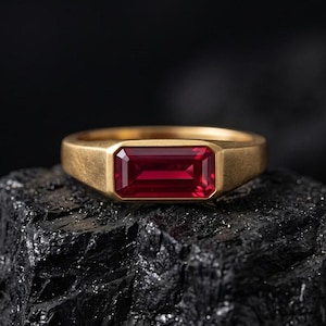 Könnte beinhalten: Ein goldfarbener Ring mit einem rechteckigen, tiefroten Edelstein. Der Ring befindet sich auf einer dunklen, strukturierten Oberfläche. Der Edelstein steht im Mittelpunkt, das Band hat ein schlichtes, klassisches Design.