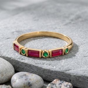 Puede incluir: Un anillo de banda dorado con piedras preciosas rojas rectangulares y verdes redondas alternadas. El anillo se exhibe sobre una superficie de piedra gris, resaltando el diseño y el contraste de color de la joya.