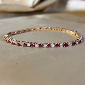 Könnte beinhalten: Ein goldenes Tennisarmband mit einem abwechselnden Muster aus rund geschliffenen, tiefroten Rubinen und klaren, funkelnden weißen Diamanten in einer durchgehenden Reihe. Das polierte Goldband zeigt ein klassisches Design mit sicherem Verschluss.