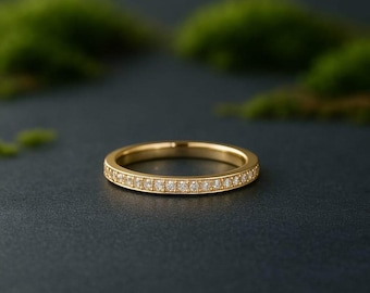 Può includere: Un anello a fascia in oro con una fila di piccole pietre preziose trasparenti incastonate attorno alla fascia. L'anello è su una superficie scura con un fogliame verde sfocato sullo sfondo. L'anello è un gioiello delicato.