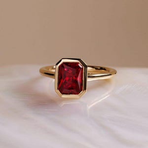 May include: A gold ring featuring a rectangular, deep red gemstone. The gemstone is set within a gold bezel, and the band is a simple, polished gold. The ring is displayed on a white, textured surface.