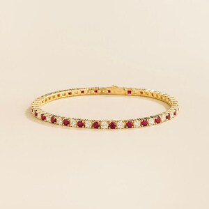 Könnte beinhalten: Ein goldenes Armband mit abwechselnd roten und weißen Edelsteinen. Das Tennisarmband-Design hat eng aneinander liegende Steine. Der Verschluss ist oben sichtbar. Der Hintergrund ist einfarbig und neutral.