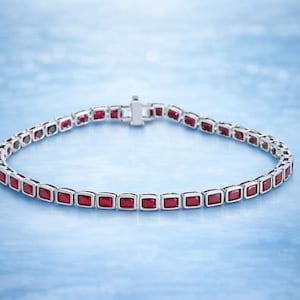 Peut inclure: Un bracelet en argent orné de pierres rouges carrées. Le bracelet est présenté sur un fond bleu clair. Le fermoir est visible, et le design est élégant et classique. Les pierres sont espacées uniformément.