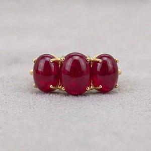 Puede incluir: Un anillo de oro con tres piedras preciosas ovaladas de color rojo intenso. Las piedras están engastadas en una banda de oro, creando un diseño clásico y elegante. El anillo se muestra sobre un fondo gris neutro.