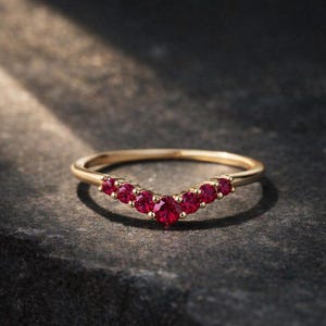 May include: A gold ring featuring a curved row of bright red gemstones. The ring's slender band supports the gemstones, which are evenly spaced. The ring is presented on a dark, textured surface, highlighting the ring's design.
