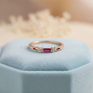 Könnte beinhalten: Ein roségoldener Ring mit einem rechteckigen Rubin-Mittelstein und kleinen Diamant-Akzenten. Der Ring liegt auf einer hellblauen Oberfläche. Der Ring ist ein Schmuckstück.