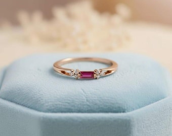 May include: A rose gold ring with a rectangular ruby center stone and small accent diamonds. The ring is set on a light blue surface, showcasing its elegant design. The ring is a piece of jewelry.