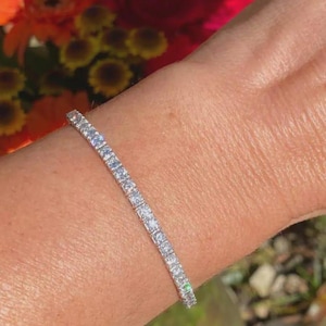 Könnte beinhalten: Ein silberfarbenes Tennisarmband mit klaren Edelsteinen. Das Armband wird am Handgelenk getragen. Der Hintergrund ist verschwommen und zeigt orange und rote Blumen. Das Armband ist etwa 18 cm lang.
