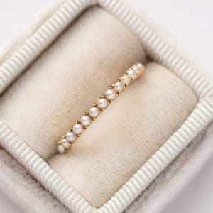 Op de afbeelding: Een delicate gouden ring bezet met een rij kleine, ronde parels. De ring wordt gepresenteerd in een zachte, crèmekleurige sieradendoos, wat een contrast creëert tussen het warme goud en de koele, neutrale achtergrond. De parels zijn gelijkmatig verdeeld.
