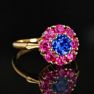 Könnte beinhalten: Ein goldener Ring mit einem zentralen blauen Edelstein, umgeben von rosa Edelsteinen. Der Ring ist vor einem schwarzen Hintergrund platziert, mit einer Spiegelung des Rings darunter.