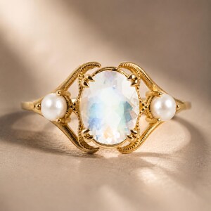 Könnte beinhalten: Ein Goldring mit einem zentralen, oval geschliffenen, schillernden Mondstein in einer Krappenfassung. Der Ring ist mit zwei kleinen, runden, weißen Perlen verziert, die sich auf beiden Seiten des Mittelsteins in einer verzierten Goldfassung befinden.
