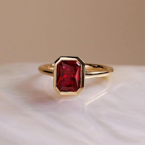 Könnte beinhalten: Ein goldener Ring mit einem rechteckigen, tiefroten Edelstein. Der Edelstein ist in einer goldenen Lünette gefasst, und das Band ist aus poliertem Gold. Der Ring wird auf einer weißen, strukturierten Oberfläche gezeigt.