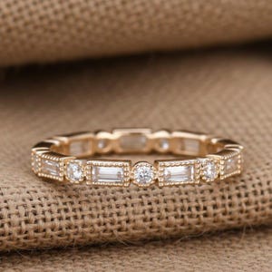 May include: A gold band ring featuring an alternating pattern of baguette-cut and round brilliant-cut clear stones. The metal setting displays a delicate beaded texture along the edges, creating a continuous design around the entire circumference.