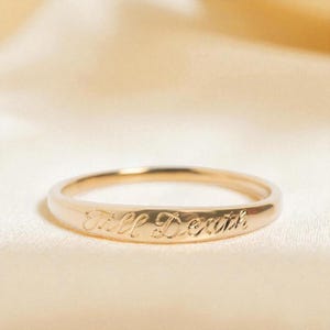 Könnte beinhalten: Ein goldfarbener Ring mit der Gravur "Till Death" auf dem Band. Der Ring, mit schlichtem Design, wird vor einem weichen, neutralen Hintergrund präsentiert, der die Eleganz der Schrift hervorhebt.