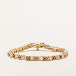 Könnte beinhalten: Ein goldenes Armband mit abwechselnd quadratisch geschliffenen roten und klaren Edelsteinen. Das Armband ist an einer Goldkette befestigt und hat einen Verschluss. Die Edelsteine sind gleichmäßig verteilt.