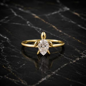 May include: A gold ring featuring a pear-shaped clear gemstone set in the center. The metal setting is shaped like a sea turtle, with the pear-shaped stone forming the shell and four small gold prongs extending outward to resemble turtle flippers.