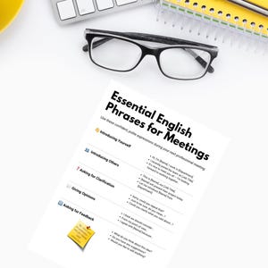 Op de afbeelding: Een werkplek met een toetsenbord, een bril en een notitieblok. Een afgedrukt vel met de titel "Essential English Phrases for Meetings" staat centraal en biedt advies voor professionele communicatie.
