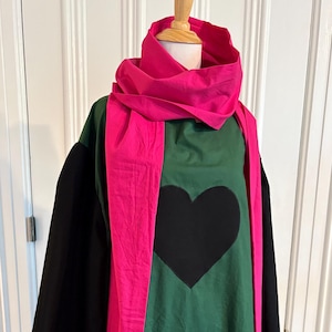 May include: A long-sleeved garment featuring a forest green body with a large black heart centered on the chest. The piece includes wide black sleeves and a bright fuchsia pink scarf draped around the neck, extending down the front of the torso.