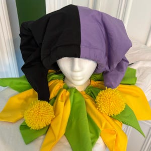 May include: A jester hat featuring a split black and purple design, paired with a ruffled collar in alternating yellow and lime green fabric. The collar is accented with two bright yellow pom-poms, creating a vibrant, multi-colored costume accessory.