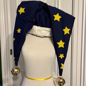 Könnte beinhalten: Eine blaue und gelbe Narrenmütze mit goldenen Glöckchen. Die Mütze hat eine spitze Krone und lange, fließende Spitzen. Die Mütze ist mit gelben Sternen verziert.