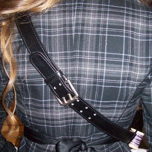 May include: A black and white plaid jacket with a black leather strap with silver hardware. The strap has white stitching and is worn diagonally across the back of the jacket.
