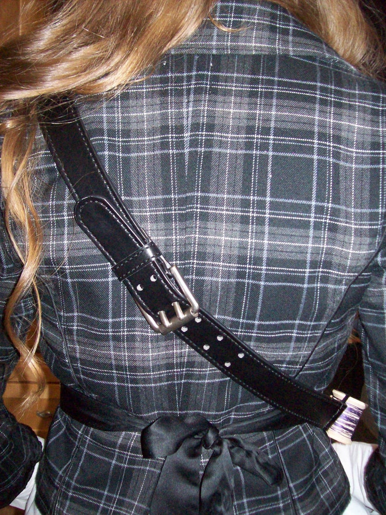 May include: A black and white plaid jacket with a black leather strap with silver hardware. The strap has white stitching and is worn diagonally across the back of the jacket.